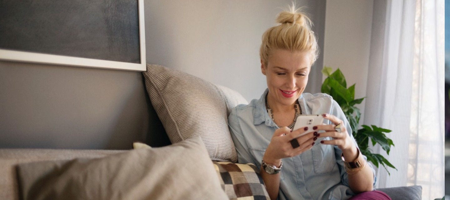 A woman is seated on a couch in her home smiling at her phone.