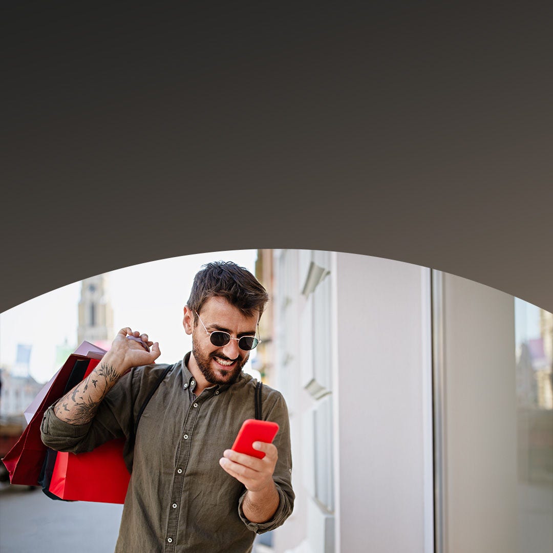 Man smiling as he uses his smartphone, holding shopping bags in his other hand.